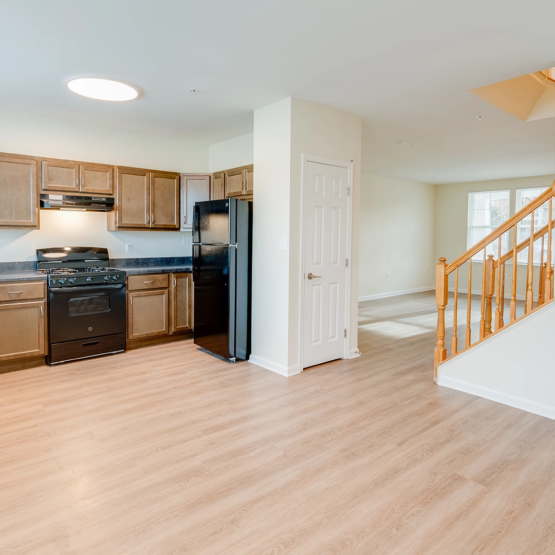 Modern kitchen with brown cabinetry, black appliances, and hardwood flooring, leading to a bright open living space with a staircase.